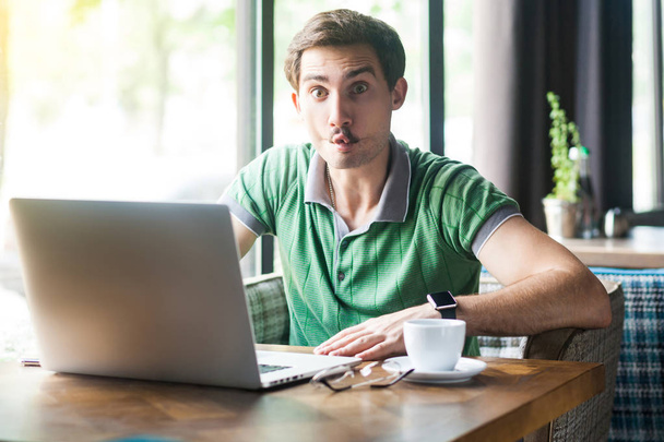 Young funny businessman in green t-shirt sitting in cafe and working on laptop while looking at camera and showing fish lips gesture. business and freelancing concept - Photo, Image