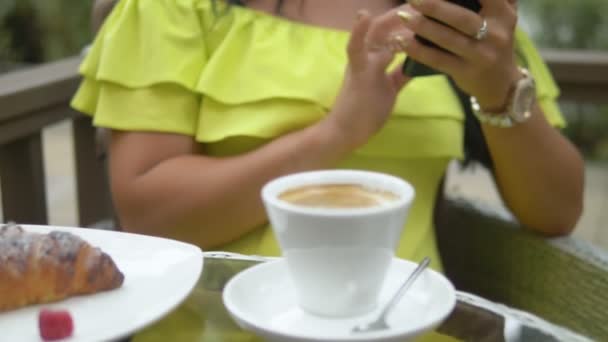 Beautiful brunette woman using cellphone at coffee shop outdoors. Breakfast with a croissant and coffee, checking the news on your smartphone. - Footage, Video