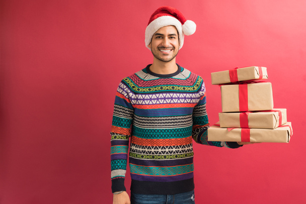 Handsome young Hispanic man wearing Christmas hat and ugly sweater while holding presents against red background - Φωτογραφία, εικόνα