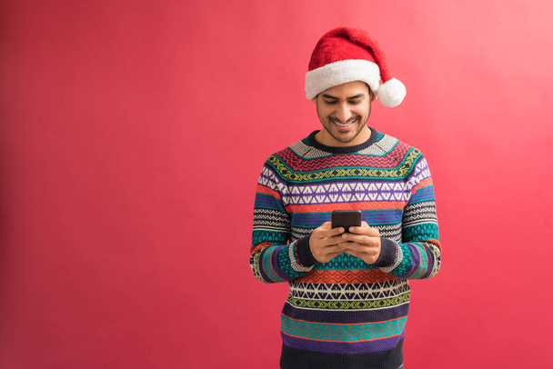 Happy Latin man text messaging on smartphone while celebrating Xmas against plain background - Photo, Image