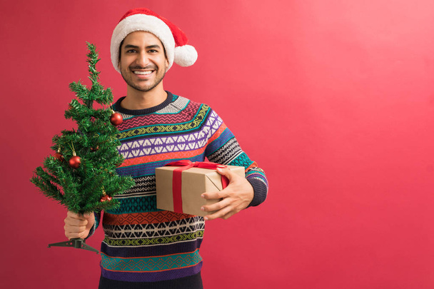Handsome young Hispanic man holding Christmas tree and present against plain background - Photo, Image