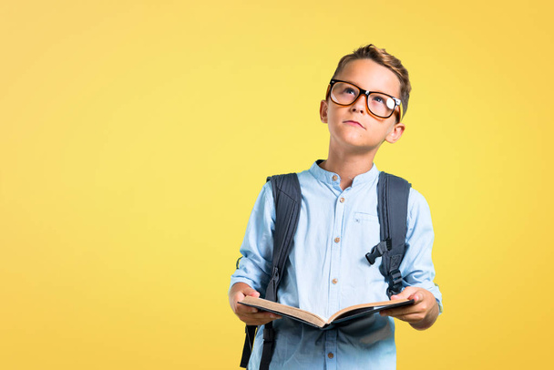 Студент в рюкзаке и очках держит книгу на желтом фоне
 - Фото, изображение