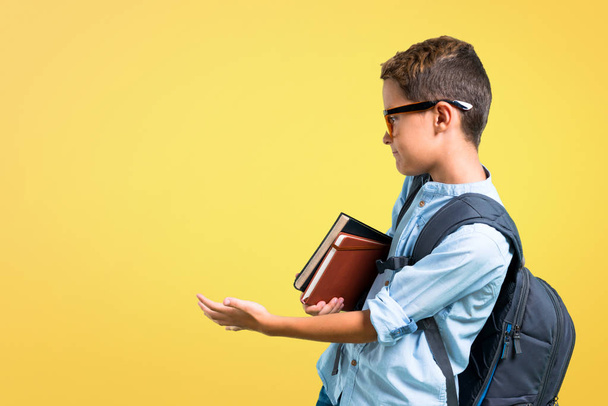 Μαθητής αγόρι με σακίδιο και γυαλιά που δείχνουν πίσω με το δείκτη του χεριού σε κίτρινο φόντο - Φωτογραφία, εικόνα