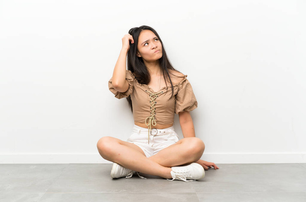 Teenager asian girl sitting on the floor having doubts and with confuse face expression - Foto, Imagen