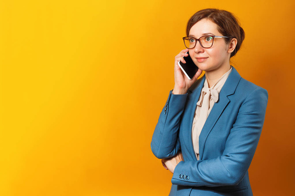 Young business woman in glasses and a suit talking on a cell phone on a yellow background. - Фото, изображение