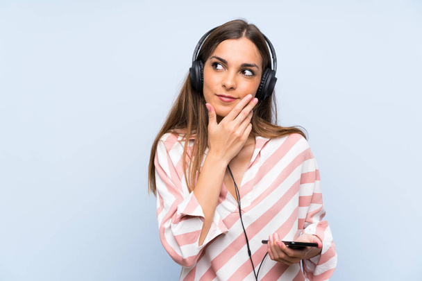 Mladá žena naslouchající hudbě s mobilním přes izolovanou modrou zeď, která uvažuje o myšlence - Fotografie, Obrázek