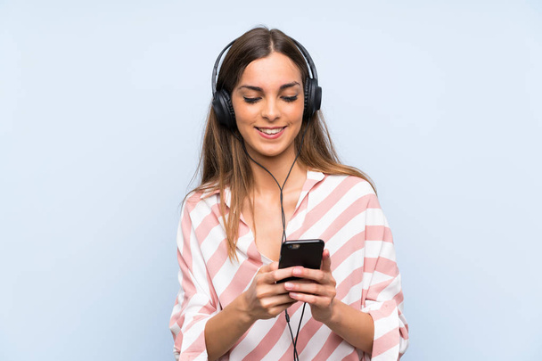Fiatal nő zenét hallgat egy mobil felett elszigetelt kék fal - Fotó, kép