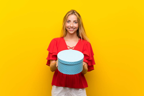 Blonde young woman over isolated yellow background holding gift box - Fotografie, Obrázek