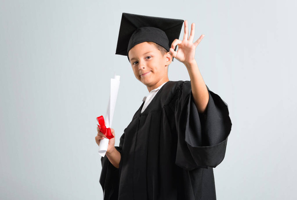 Kleine jongen afstuderen het tonen van een OK teken met vingers op grijze achtergrond - Foto, afbeelding