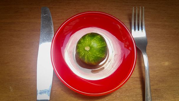 un piccolo cetriolo su un piatto rosso, ma il cetriolo è molto simile a un piccolo cocomero o pianeta Terra, accanto a un coltello e una forchetta, come se la gente avida volesse divorare il pianeta terra
 - Foto, immagini