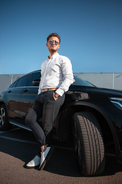 Jonge succesvolle zakenman in een wit shirt en zonnebril staan in de buurt van zijn luxueuze zwarte auto. - Foto, afbeelding