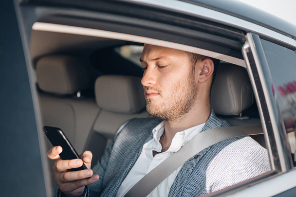 Portrait of a young businessman in a suit on a rear seat checking news with smartphone. - Φωτογραφία, εικόνα