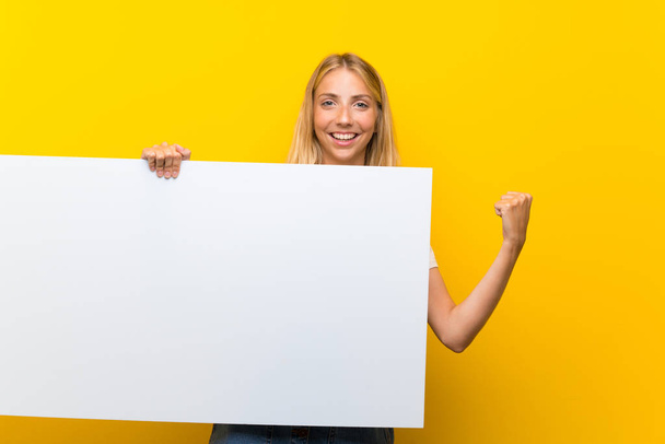 Blonde young woman over isolated yellow background holding an empty white placard for insert a concept - Photo, Image