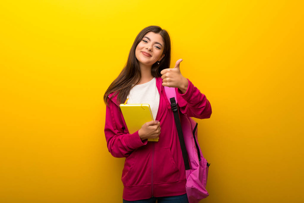 Adolescente estudante menina no fundo amarelo vibrante dando um gesto polegar para cima e sorrindo
 - Foto, Imagem