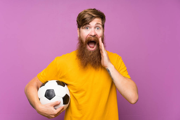 Redhead s dlouhým plnovousem přidržuje fotbalový míč nad osamocenou purpurovou stěnou s překvapením a šokovaným výrazem obličeje - Fotografie, Obrázek