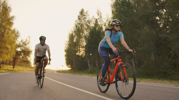 Két kerékpáros egy férfi és egy nő lovagolni az autópályán a közúti kerékpárok viselése sisakok és sportruházat a naplemente lassított. - Felvétel, videó