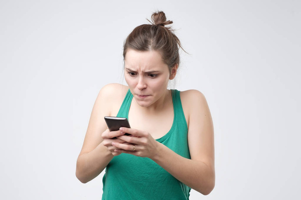 Młoda kaukaski kobieta w zielonej koszulce patrząc na telefon z podrażnieniem. - Zdjęcie, obraz