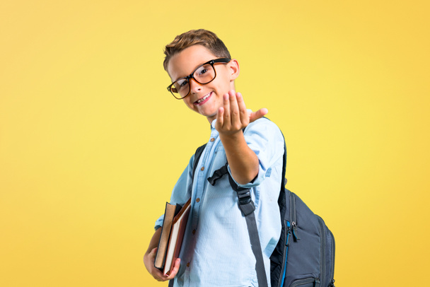Student boy with backpack and glasses presenting and inviting to come on yellow background - Fotografie, Obrázek