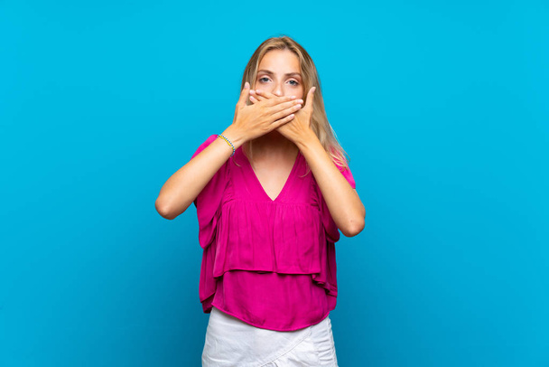 Блондинка молода жінка над ізольованим синім тлом покриває рот руками
 - Фото, зображення