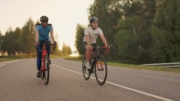 Twee fietsers een man en een vrouw rijden op de snelweg op de weg fietsen dragen helmen en sportkleding bij zonsondergang in slow motion. - Video