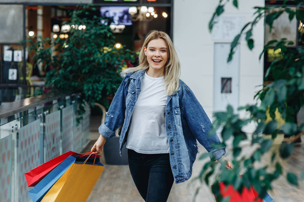 Beautiful blonde girl in a denim jacket with bags at the shopping center. She is carrying shopping bags after shopping and looking at camera. Model in sales, shop, retail, consumer concept - Photo, image