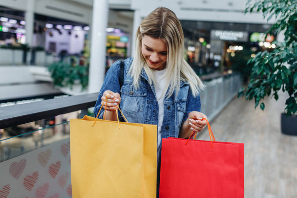 買い物の季節。ぼやけたショッピングモールの背景にカラフルなショッピングバッグとデニムジャケットの美しい女の子。買い物袋の中を見ている女性。左側または右側の領域をコピーする - 写真・画像