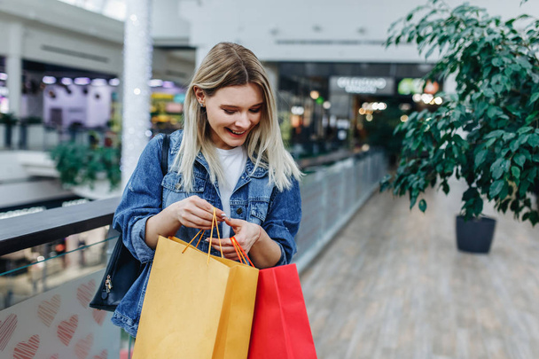 Shopping season. Beautiful laughing girl in a denim jacket with bags on blurred shopping mall background. Urban life and shopogolic concept. Copy space on the right side - Φωτογραφία, εικόνα