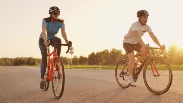 İki bisikletçi bir adam ve bir kadın yavaş hareket gün batımında kask ve spor giyim giyen yol bisikletleri üzerinde yolculuk. - Video, Çekim