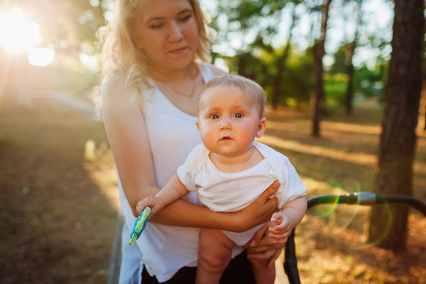 Madre joven caminando en el parque de verano con su bebé. Mujer feliz sosteniendo niño al aire libre
 - Foto, Imagen
