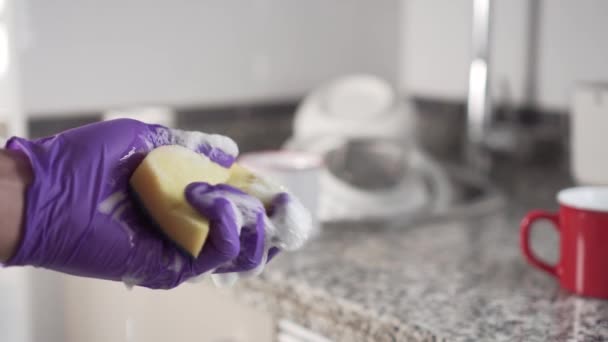 Une main dans un gant de protection bleu serre une éponge jaune-vert mousseuse sur un fond de montagne de vaisselle non lavée dans la cuisine de l'évier
. - Séquence, vidéo