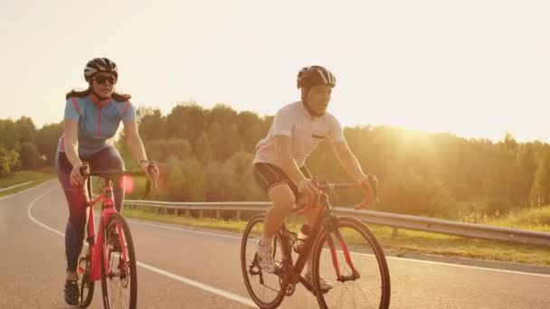 Um homem e uma mulher andam de bicicleta esportiva na estrada ao pôr-do-sol em engrenagem e capacetes de proteção em câmera lenta 120 fps
 - Filmagem, Vídeo