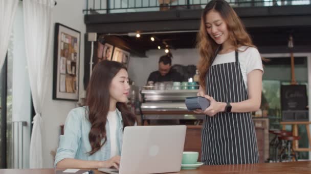 アジアの若いフリーランスの女性は、コーヒーショップで非接触クレジットカードを支払います。アジアの幸せな女の子バリスタウェイターは、顧客のためのクレジットカードリーダーマシンを保持する灰色のアポンを着用し、カフェで技術で支払うことができます. - 映像、動画