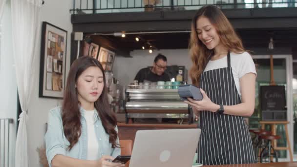 A fiatal ázsiai szabadúszó nők érintésmentesen fizetnek a kávézóban. Ázsiai boldog lány barista pincér viselni szürke kötény gazdaság hitelkártya-olvasó gép az ügyfél számára a mobiltelefon szkennelés fizetni a kávézóban. - Felvétel, videó
