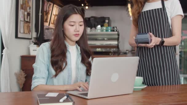 A fiatal ázsiai szabadúszó nők érintésmentesen fizetnek a kávézóban. Ázsiai boldog nő barista pincér viselni szürke kötény gazdaság hitelkártya-olvasó gép az ügyfél számára a mobiltelefon szkennelés fizetni a kávézóban. - Felvétel, videó