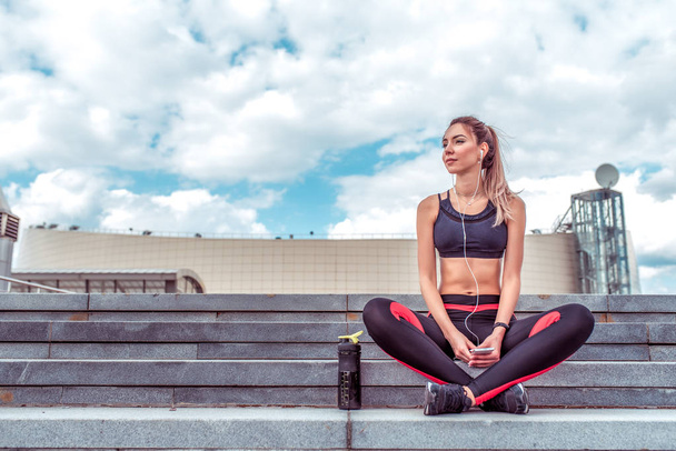 Beautiful girl athlete, in the summer in the city sits after a workout, relaxes, fitness on the street, listens to music on a smartphone, enjoy nature in the summer, a shaker with water and protein. - 写真・画像