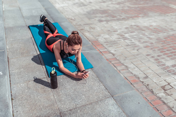 Girl athlete, lies on a yoga mat after workout, rest in city on fitness, an active lifestyle, a shaker with an energy drink and protein, headphones online application in a smartphone, free space. - Fotografie, Obrázek
