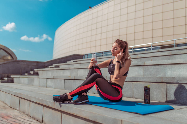 Beautiful woman sits on mat, fitness in summer city, outdoors, for yoga after workout, relaxation, active lifestyle, shaker energy drink protein, headphones online application smartphone, free space. - Photo, Image