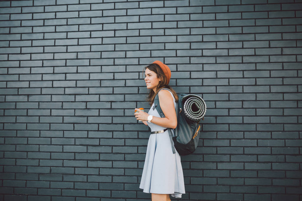 cheerful girl in dress holding take away cup. Hipster model wearing stylish summer outfit Outdoors. woman with backpack walks along city street. hipster girl on brick wall,lifestyle of modern teenage - Photo, Image