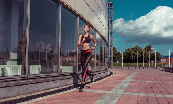 young beautiful woman jogging summer, morning in city, business lady, background glass windows building, active lifestyle, free space, sportswear leggings top figure. Clouds background, brick tile. - Photo, Image