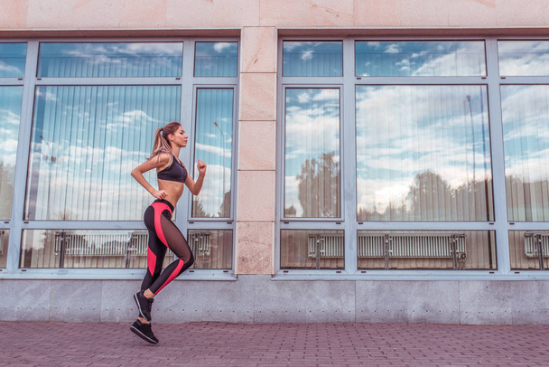 A beautiful girl in summer runs in the city on a run, the background is a building with large glass windows, an active lifestyle, free space for text, sportswear leggings top and sneakers - Photo, Image
