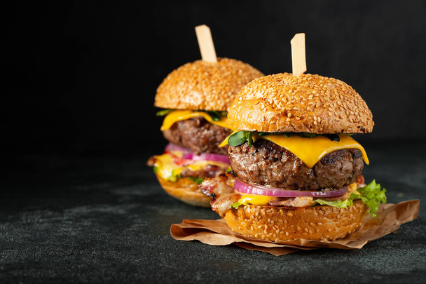 A set of homemade delicious burgers of beef, bacon, cheese, lettuce and tomatoes on a dark concrete background. Fat unhealthy food close-up. With copy space - Photo, Image