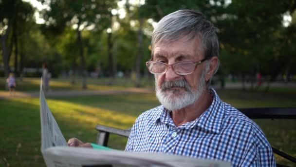 senior in plaid shirt sits on a bench in the park and reads a newspaper - Footage, Video