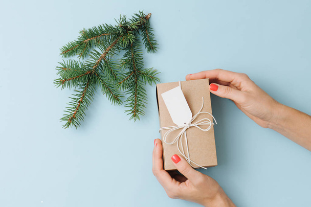 Female hands, placing gift box next to fir branch over blue background - Foto, afbeelding