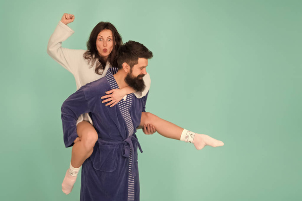 Handsome young man giving his girlfriend piggyback ride. Couple in bathrobes having fun turquoise background. Lets stay at home and have fun. They always have fun together. Close relationship - 写真・画像