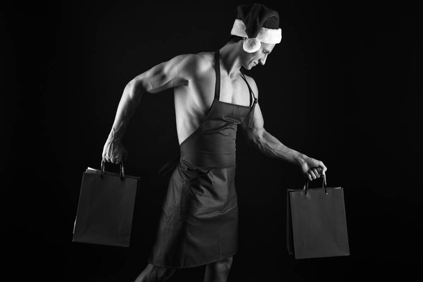 Sexy athletic macho muscular chest in santa claus hat. Athlete muscular man wear santa hat and apron. I have gift for you. Macho muscular torso posing with shopping bags. Santa claus for adult - Zdjęcie, obraz
