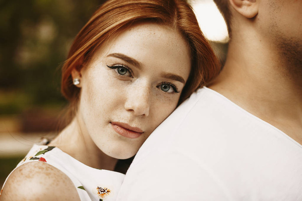 Close up portrait of a charming red hair woman with freckles and grey eyes looking at camera serious while leaning her head on the back of her boyfriend outside. - Photo, Image