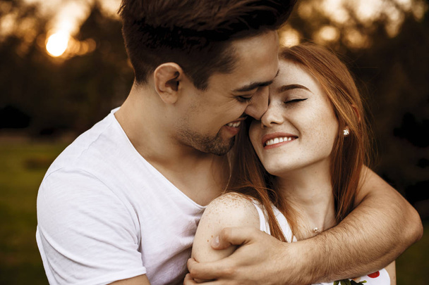 Jonge blanke lachende man omhelst van achteren zijn vriendin met sproeten en rood haar dat is met gesloten ogen glimlachen tegen zonsondergang. - Foto, afbeelding