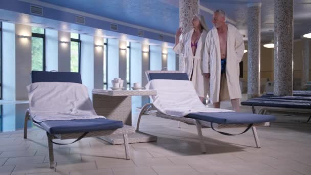 Пара пожилых людей отдыхает в отеле возле бассейна в помещении
 - Кадры, видео