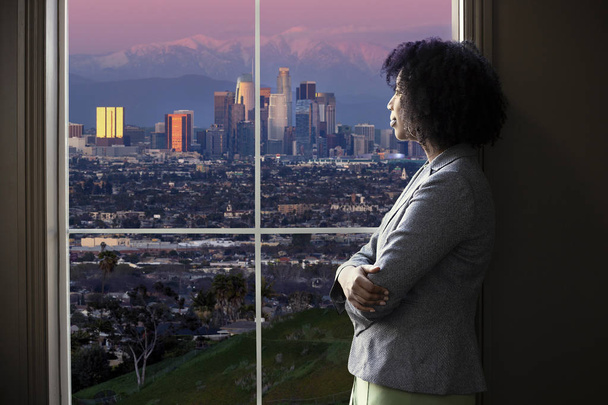 Mujer negra de negocios mirando por la ventana de una oficina en Los Ángeles. Parece una arquitecta pensando en el desarrollo urbano o un alcalde o gobernador de la ciudad planeando leyes de zonificación.
.  - Foto, Imagen