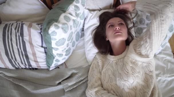 Young attractive woman in a white sweater lies on a bed, dreams and goes to bed - Footage, Video